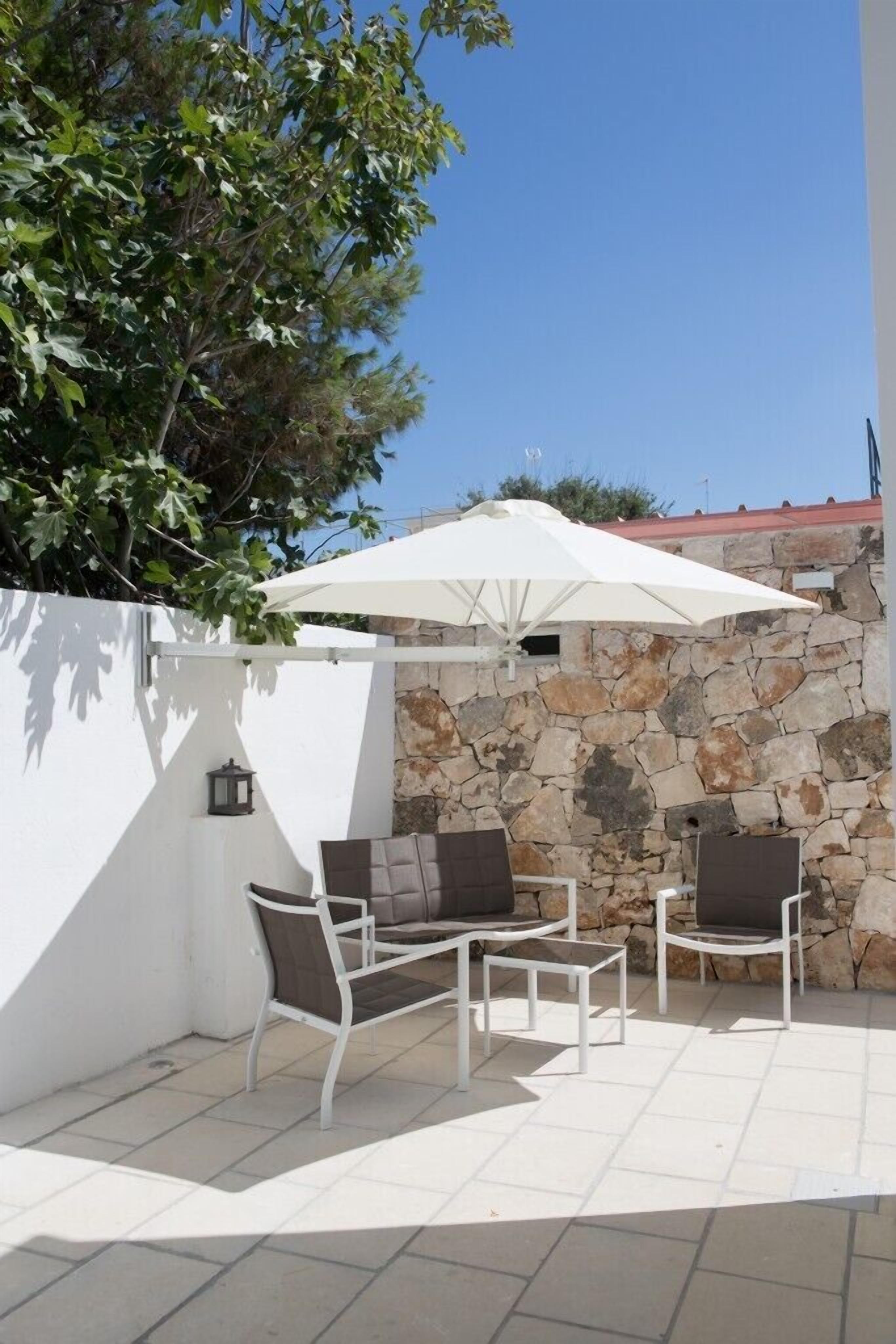An outdoor patio with white tiled flooring, a stone wall, and a white umbrella providing shade.