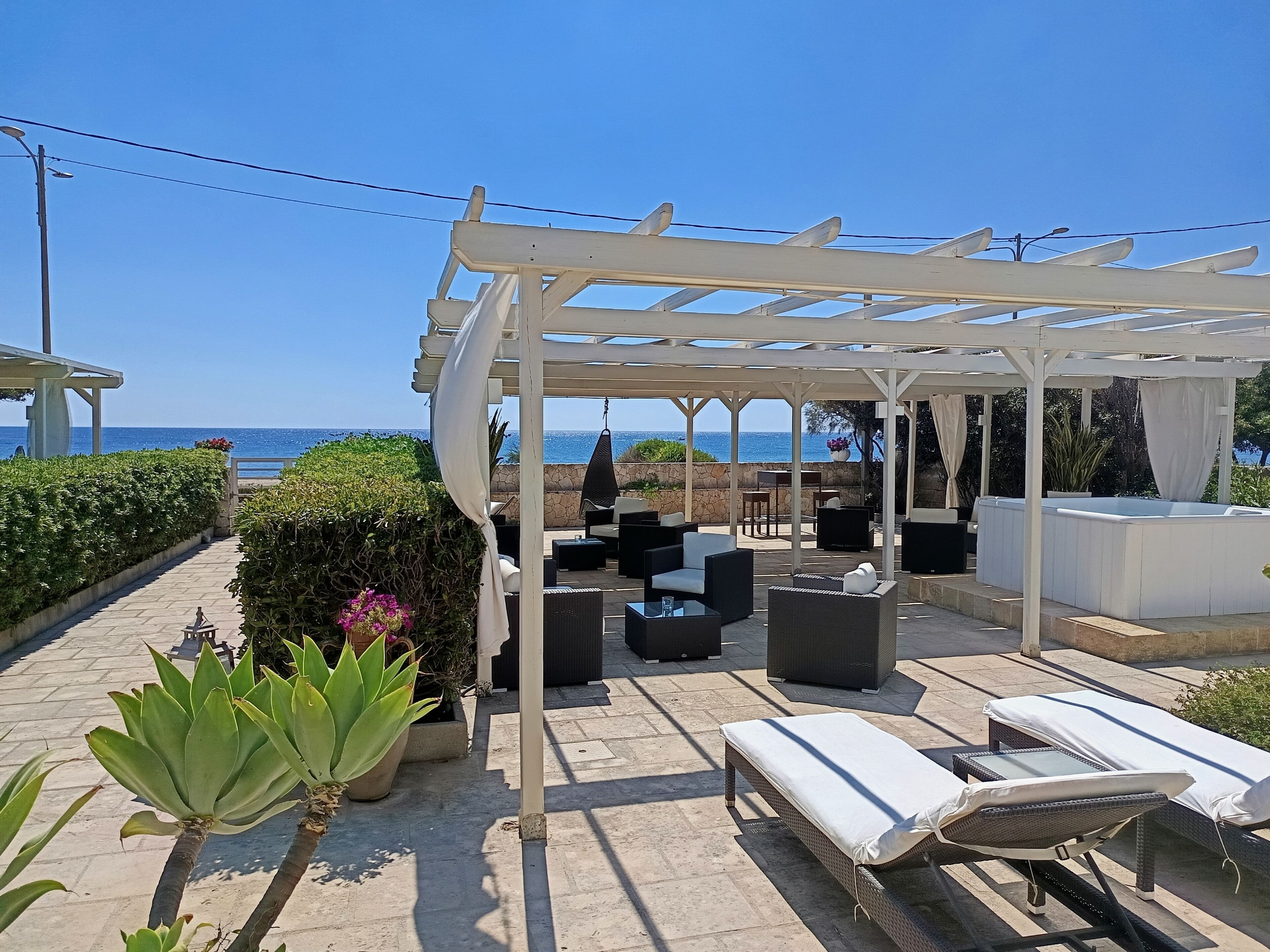 A covered outdoor seating area with lounge chairs and tables, overlooking the sea.