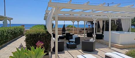 A covered outdoor seating area with lounge chairs and tables, overlooking the sea.