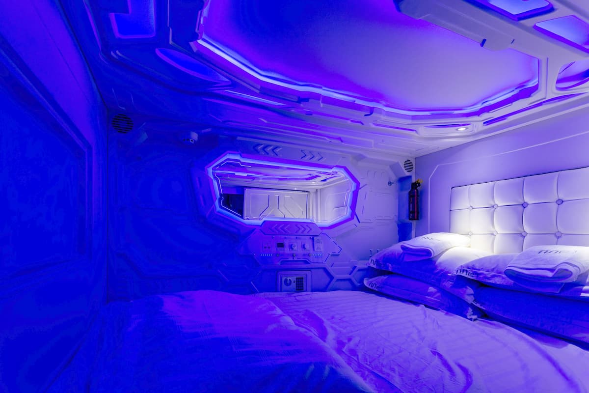 Um quarto futurista com um painel de controle central, uma cama com cabeceira estofada e uma janela com design geométrico.