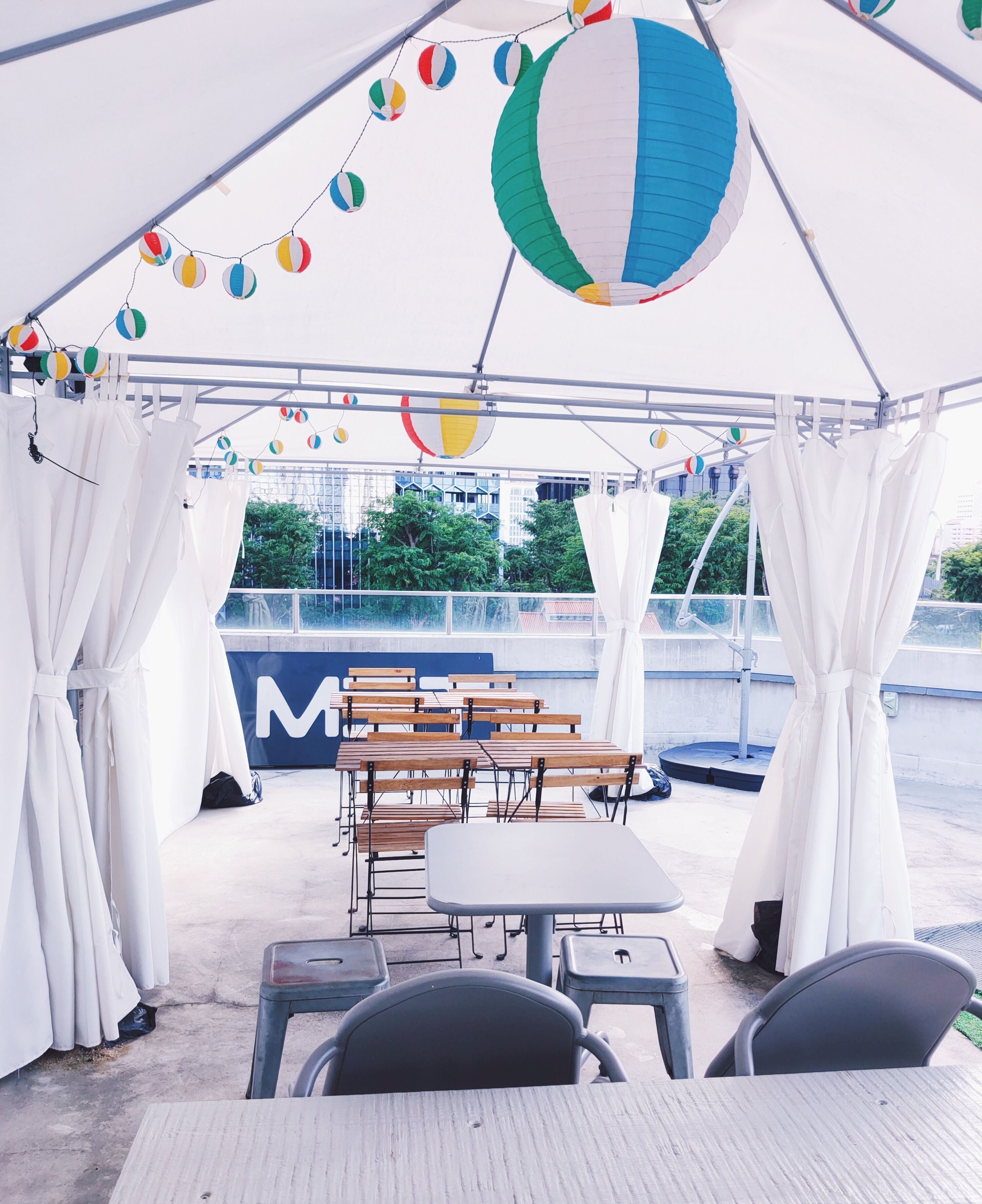 Un espacio al aire libre techado con mesas y sillas, una carpa grande y decoraciones colgantes de colores.