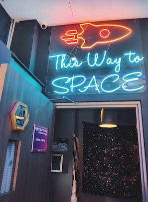 A neon sign reading "This Way to SPACE" above a reception desk, a starry night sky poster, and a reception sign indicating operating hours.