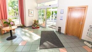 Een hotel lobby met een receptiebalie, een bank, een kleintje tafeltje met bloemen, een vloermat en een deur.