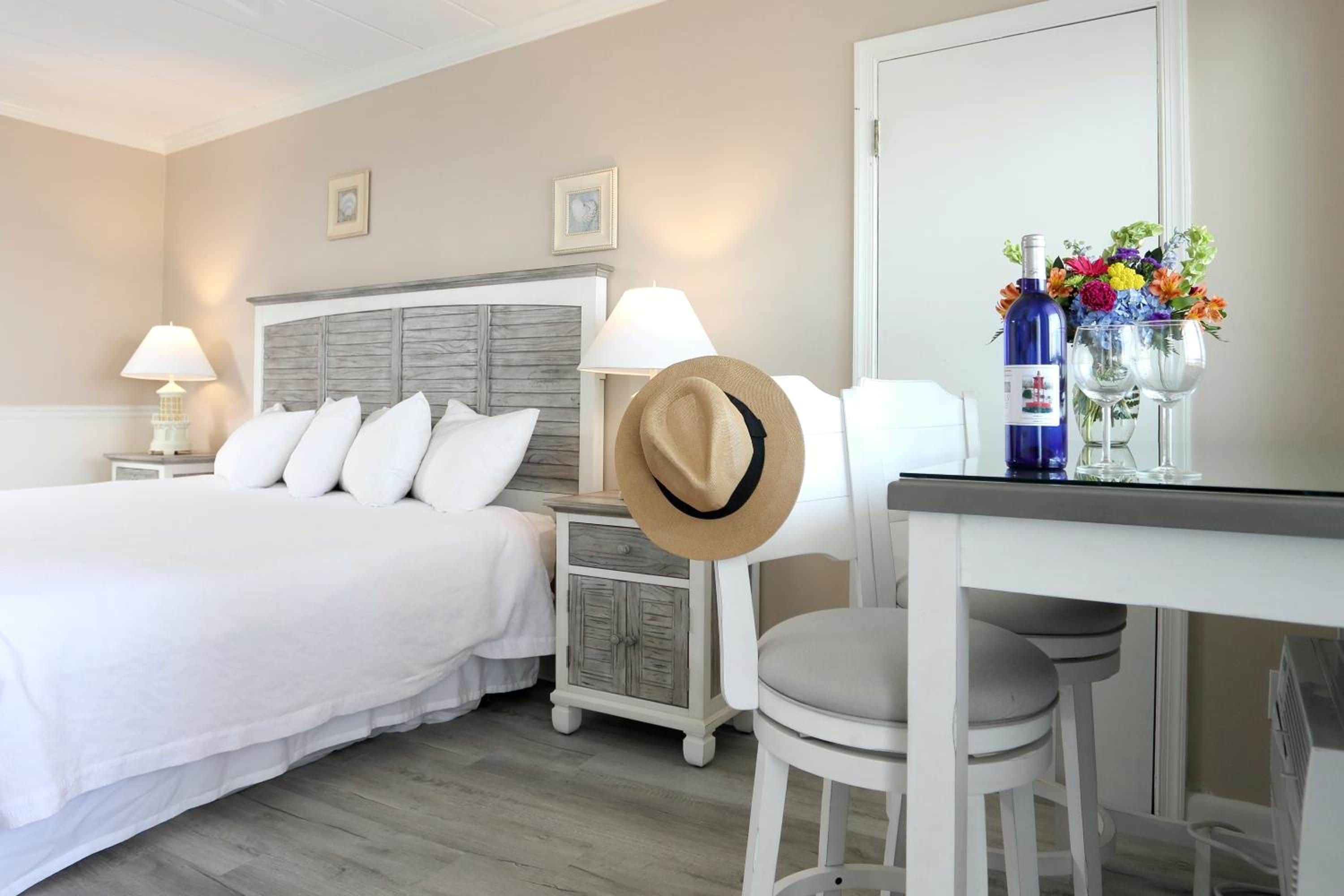 A hotel room with a bed, a desk, a chair, a lamp, a hat, and a vase with flowers.