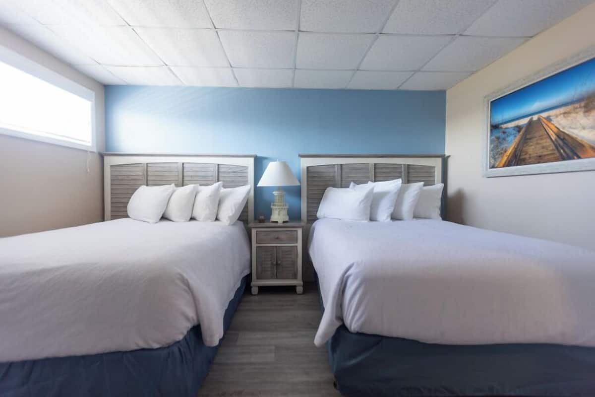 A hotel room with two beds, a nightstand with a lamp, a painting on the wall, and a window providing natural light.