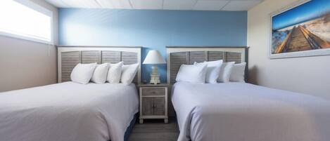 A hotel room with two beds, a nightstand with a lamp, a painting on the wall, and a window providing natural light.