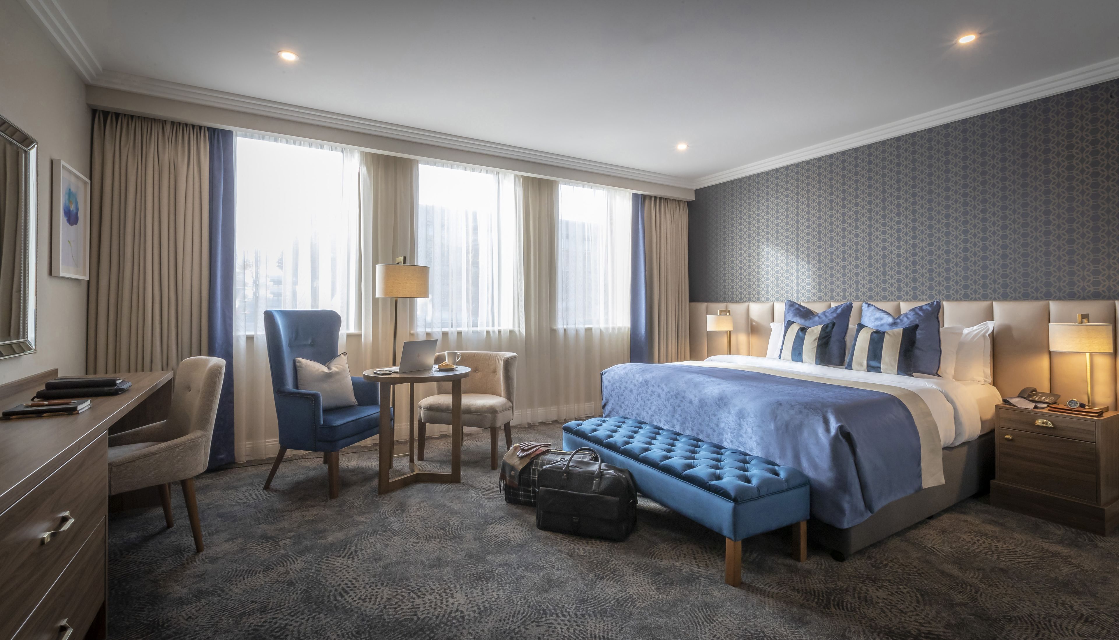 A hotel room with a large bed, a desk with a chair, a small table, and a bench.