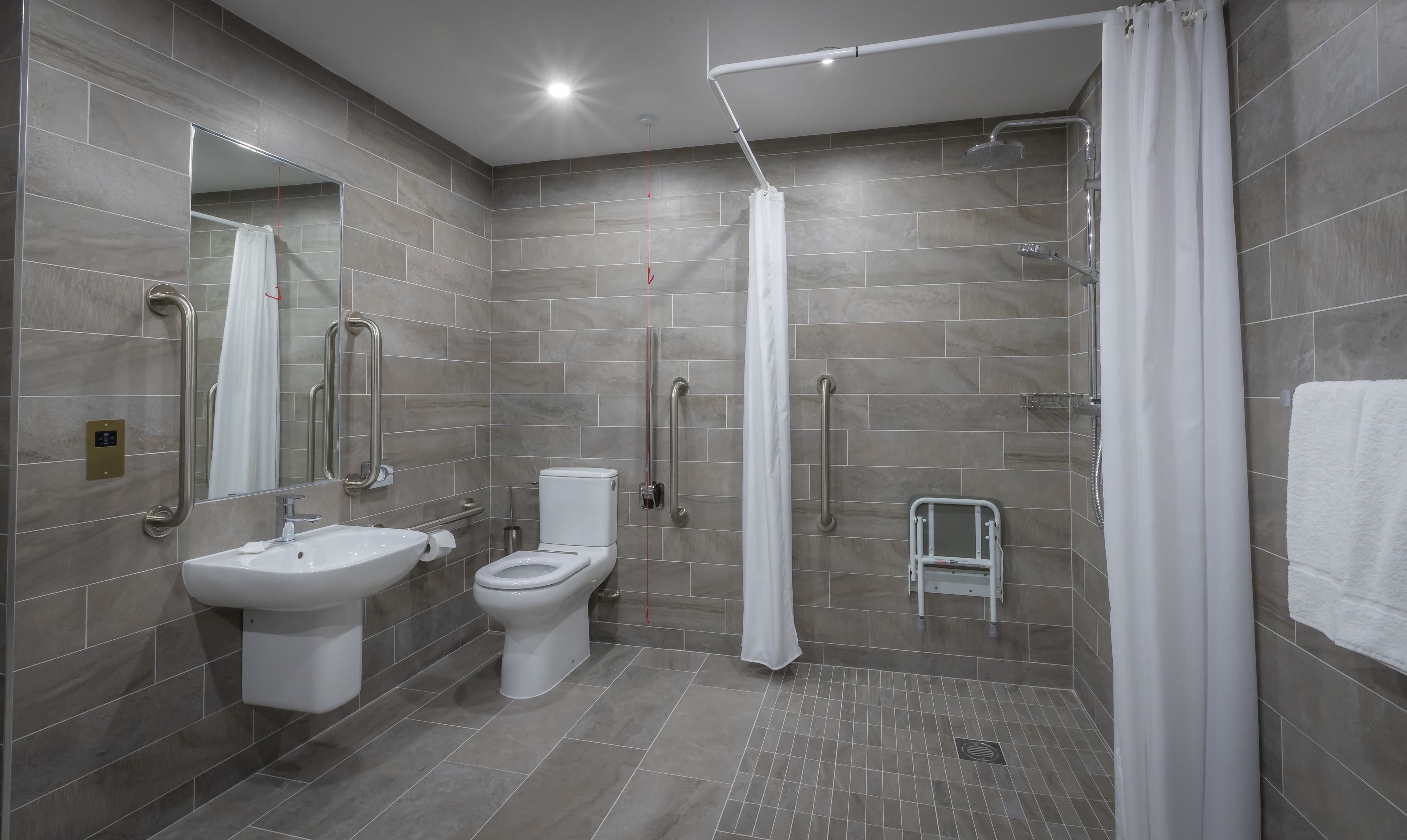 Un baño moderno con ducha, inodoro y lavamanos, que cuenta con barras de apoyo y un banco accesible para sillas de ruedas.