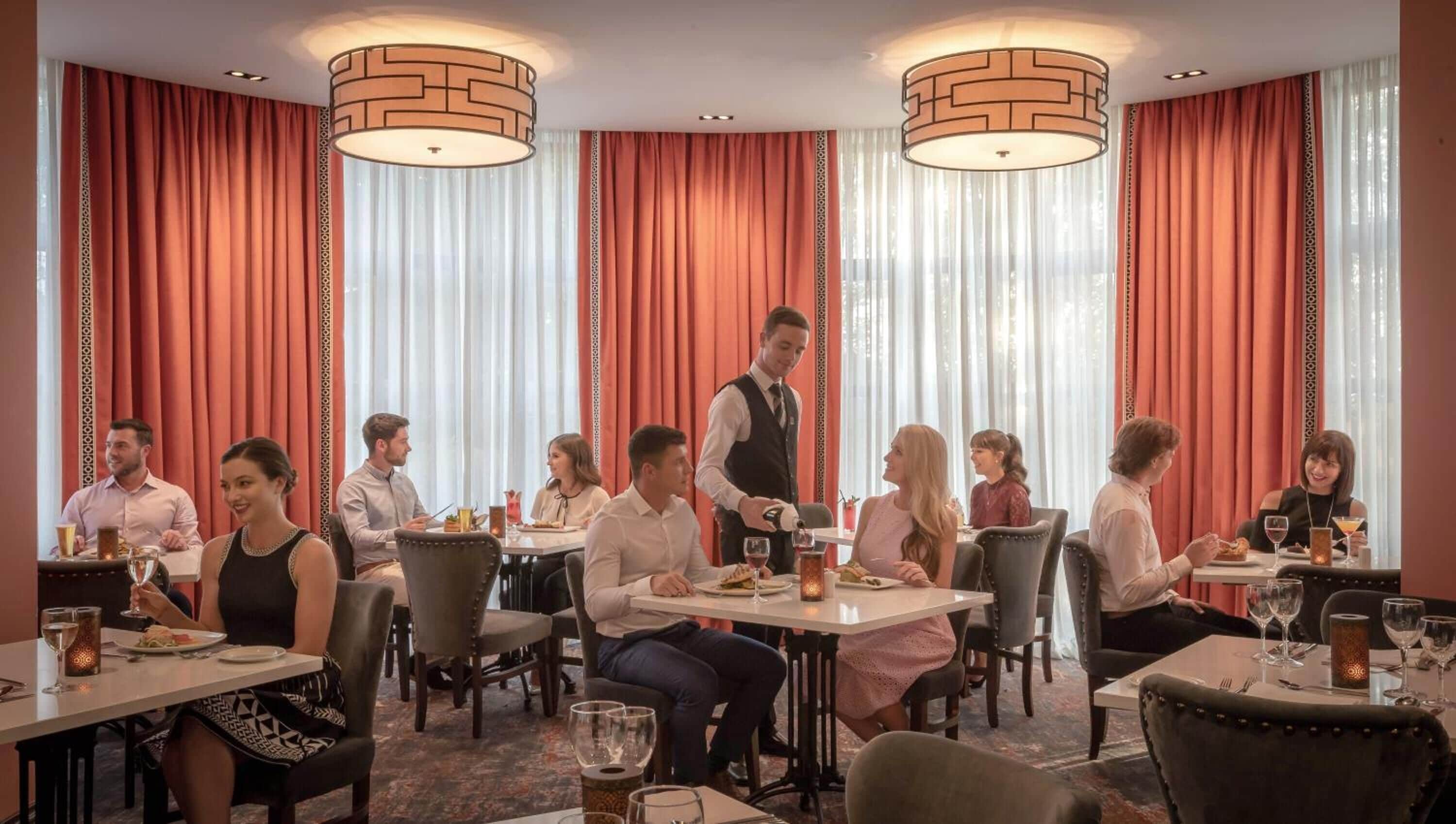 Ein Restaurant mit runden Tischen, roten Vorhängen und einem Kellner, der Getränke serviert.