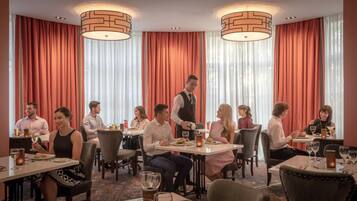 Ein Restaurant mit runden Tischen, roten Vorhängen und einem Kellner, der Getränke serviert.