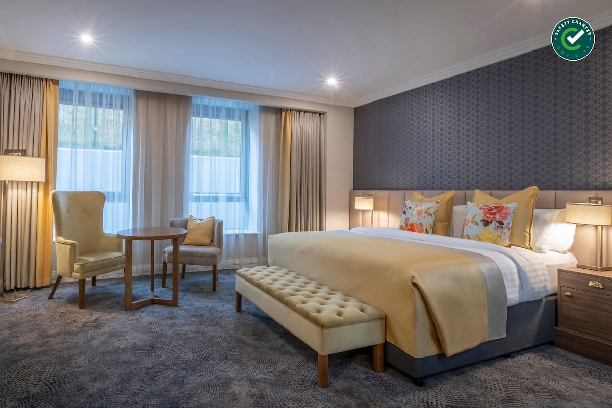 A hotel room with a large bed, a bedside table, a chair, a bench, and a window with curtains.