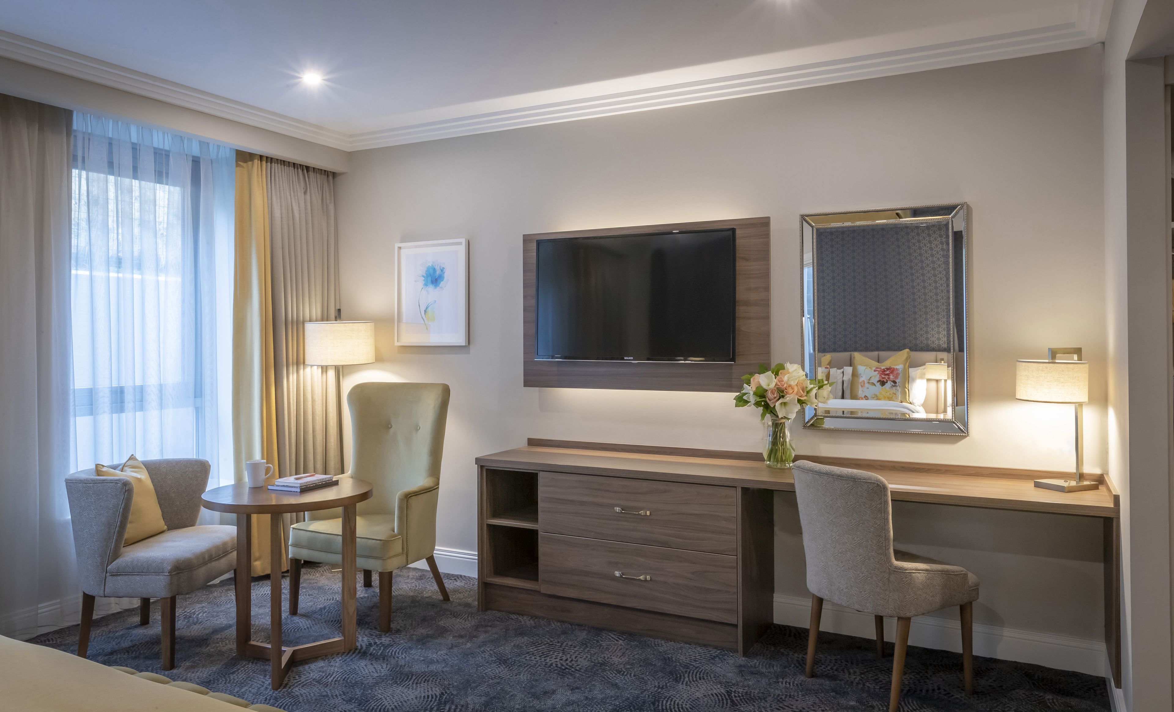 A hotel room with a desk, two chairs, a TV, a mirror, a lamp, and a vase with flowers.