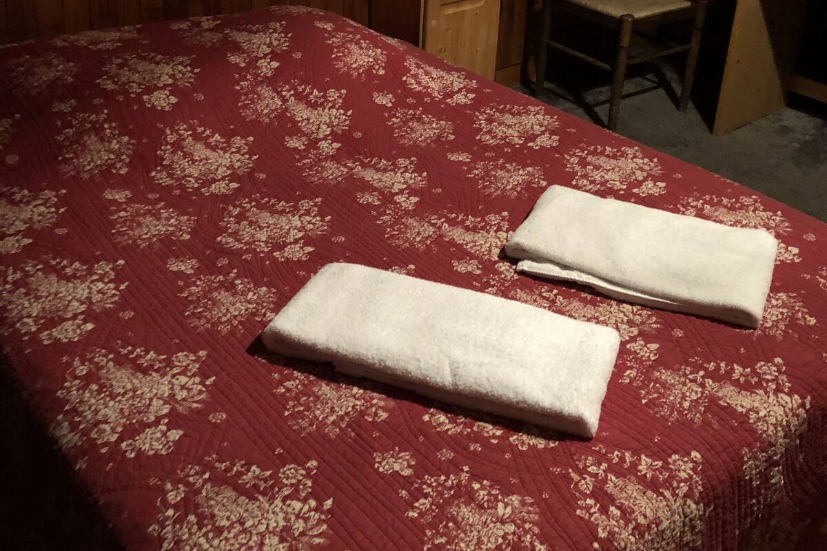 A bedroom with a wooden wall, a bed with a red floral bedspread, two folded towels, a wooden nightstand, a chair, and a desk.