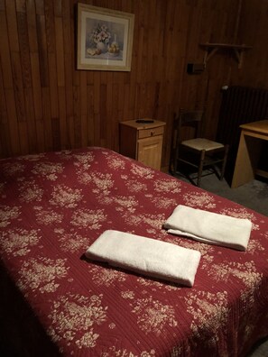 A bedroom with a wooden wall, a bed with a red floral bedspread, two folded towels, a wooden nightstand, a chair, and a desk.