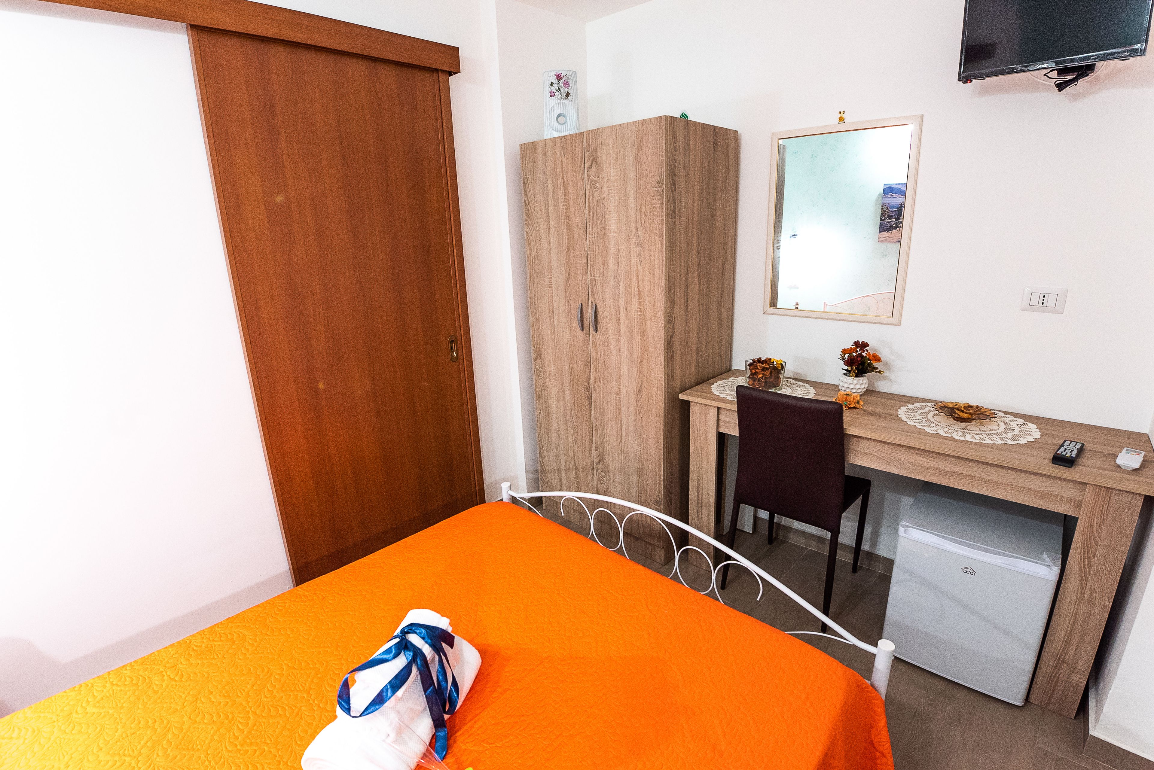 A bedroom with a bed, a desk, a chair, a wardrobe, a mirror, and a television.