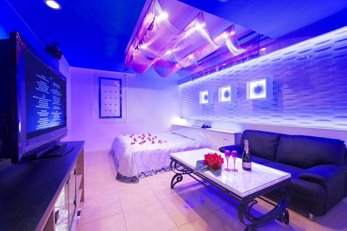 A modern room with a bed, a TV, a table with a bottle and glass, and a black sofa.