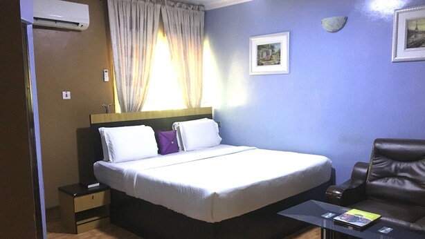 A hotel room with a bed, a chair, a nightstand, an air conditioning unit, and a window with curtains. - Sweet Savor Hotels & Suites (Lagos)
