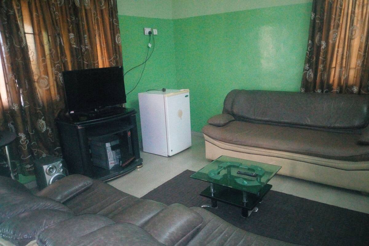 A living room with a sofa, a television on a stand, a small white refrigerator, and a glass coffee table.