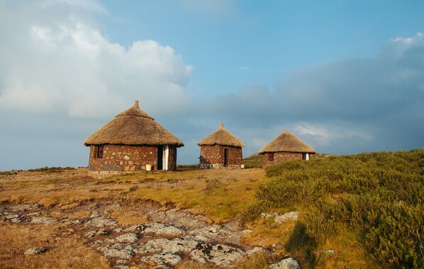 Three thatched-roof huts with stone walls, set on a grassy hill with rocky terrain.