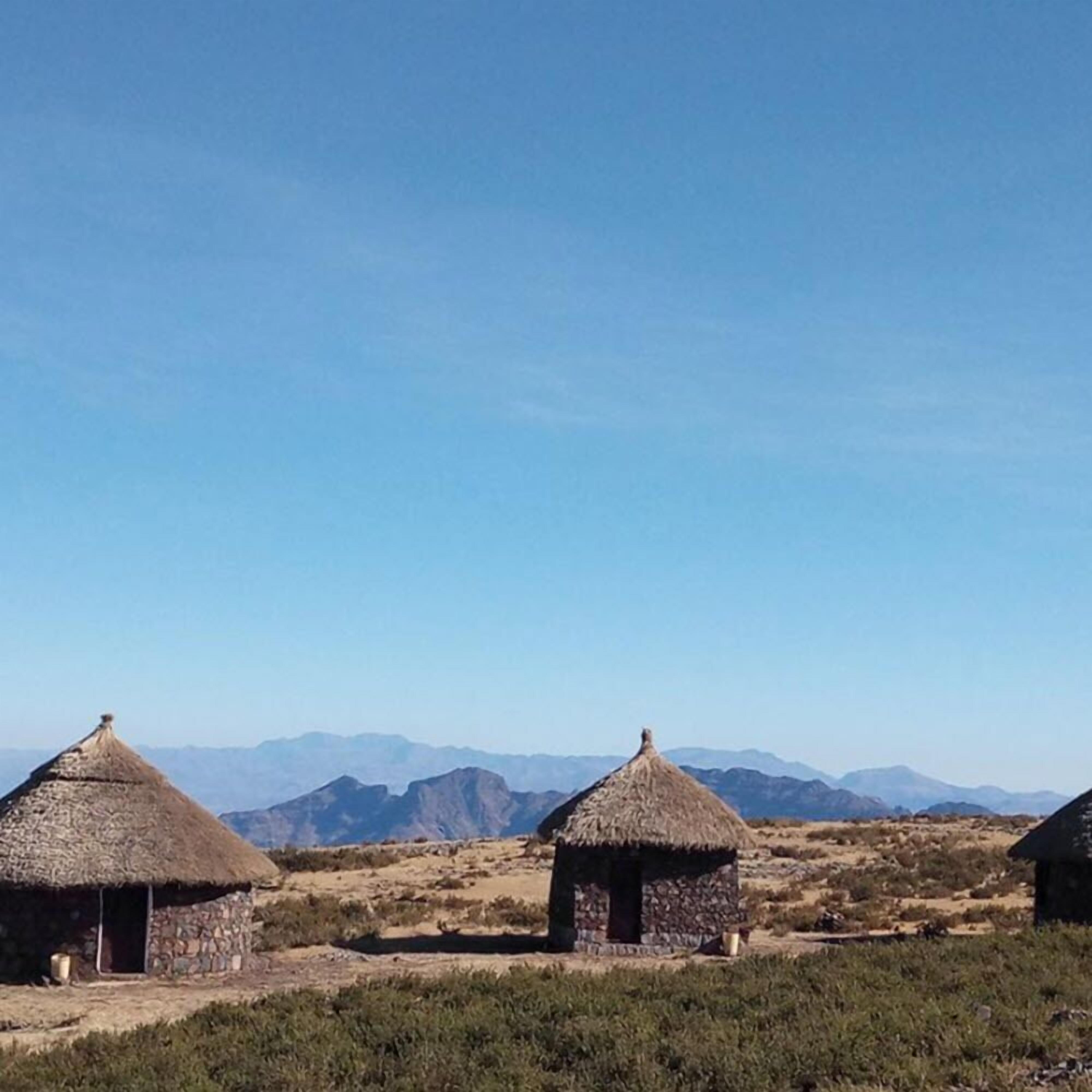 A rural landscape with thatched-roof huts and mountains in the background.