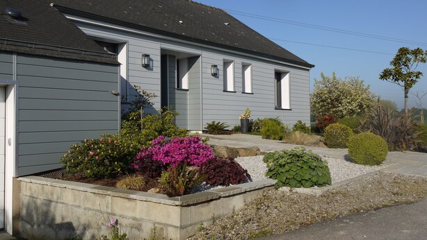 Une maison à deux étages, à l’extérieur gris, dotée d’un porche et d’un jardin aménagé avec différentes plantes et fleurs.