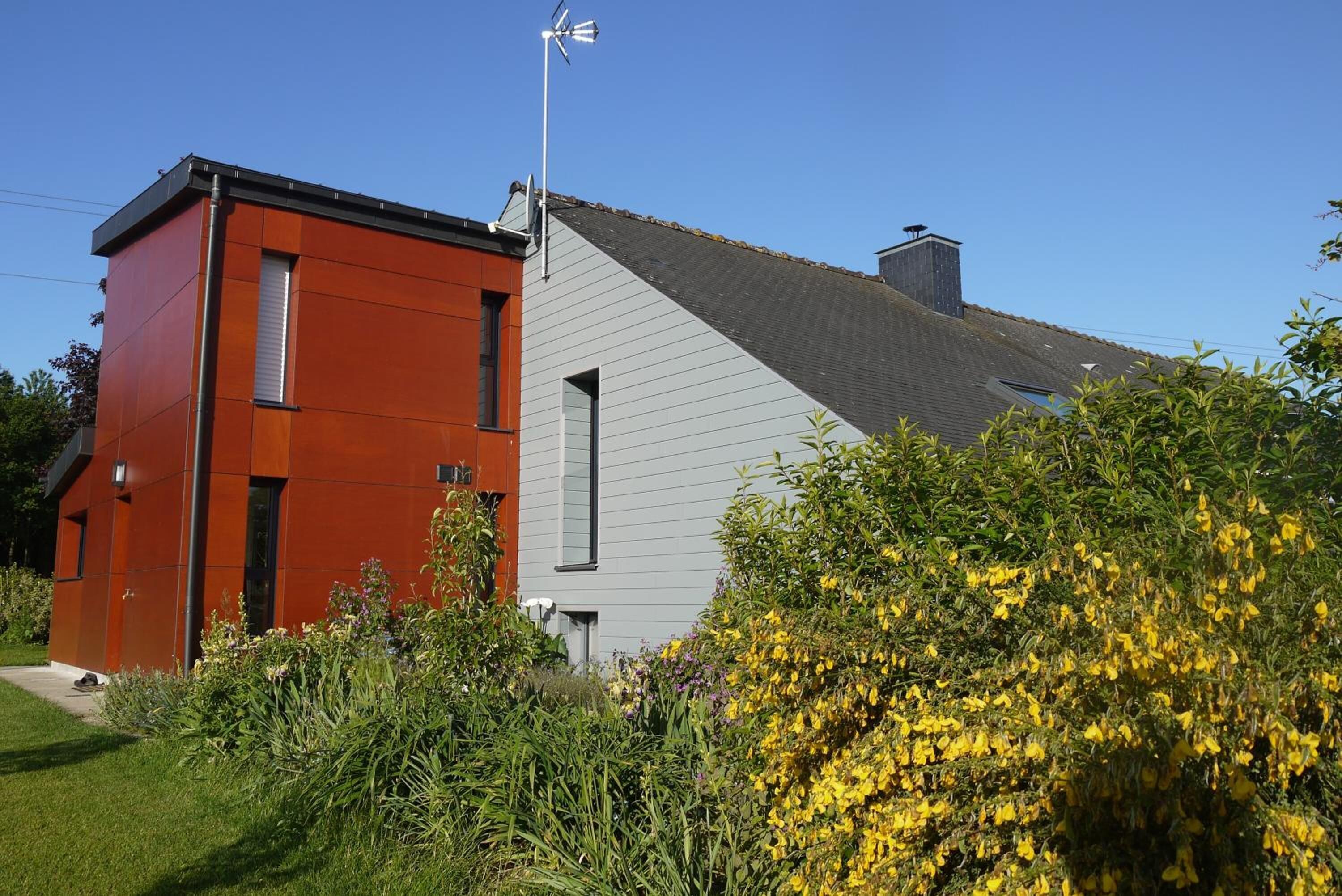 A modern two-story house with a red and white exterior, surrounded by lush greenery and yellow flowers.