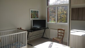 Une chambre avec un berceau, un téléviseur posé sur un meuble, une chaise et une fenêtre donnant sur des arbres.
