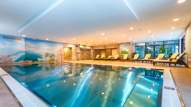 An indoor swimming pool with a mural of a mountain landscape, lounge chairs, and a large window providing a view of the outdoors. - Hotel Victoria Kaprun (Kaprun)