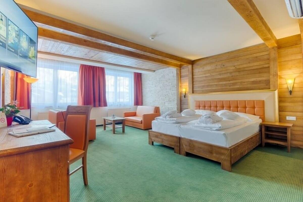 Habitación de hotel con techo de madera, cama con dos almohadas, escritorio con silla, una mesita y ventana con cortinas.