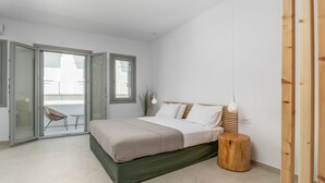 A modern bedroom with a bed, a wooden stool, and a sliding glass door leading to an outdoor area. - Olive Studios (Milos)