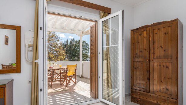 A room with a sliding glass door leading to an outdoor patio area, a wooden wardrobe, and a small table with chairs. - Olive Studios (Milos)