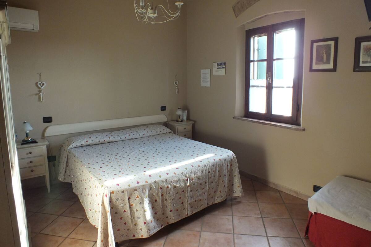 A bedroom with a bed, a nightstand, a lamp, a window, and a chandelier.