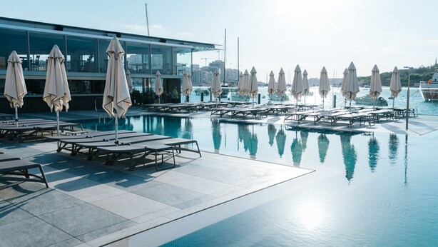 A pool area with lounge chairs and umbrellas, a modern building in the background, and a clear view of the marina with boats. - Azur Hotel by ST Hotels (Gzira)