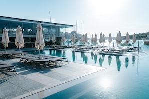 A pool area with lounge chairs and umbrellas, a modern building in the background, and a clear view of the marina with boats. - Azur Hotel by ST Hotels (Gzira)