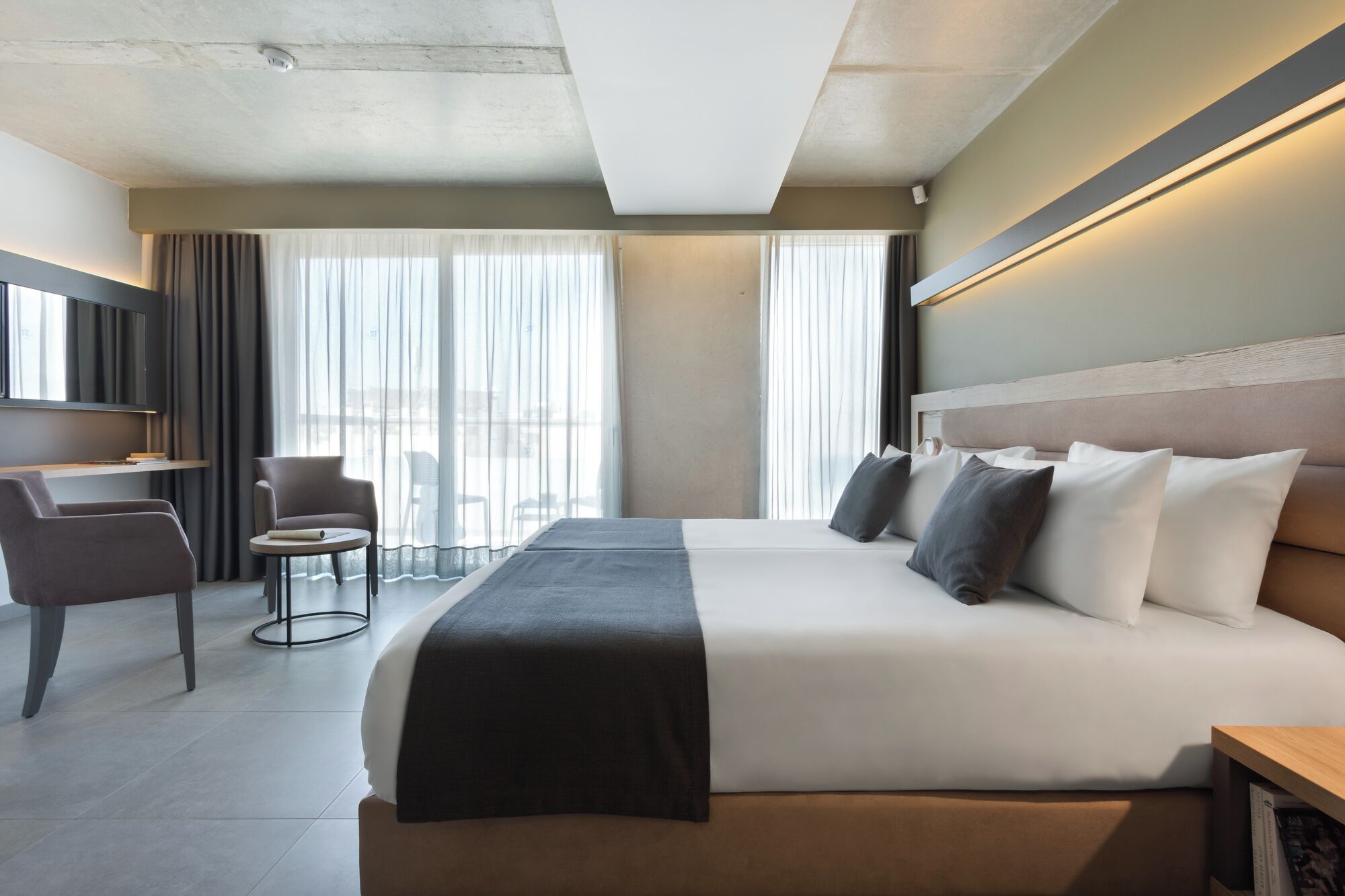 A modern hotel room with a large bed, a desk, a chair, and a view of the outdoors through large windows.