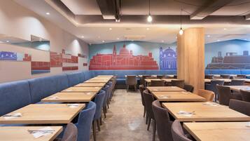 Un restaurante con largas mesas de madera y asientos tapizados en azul, con murales de paisajes urbanos en las paredes.