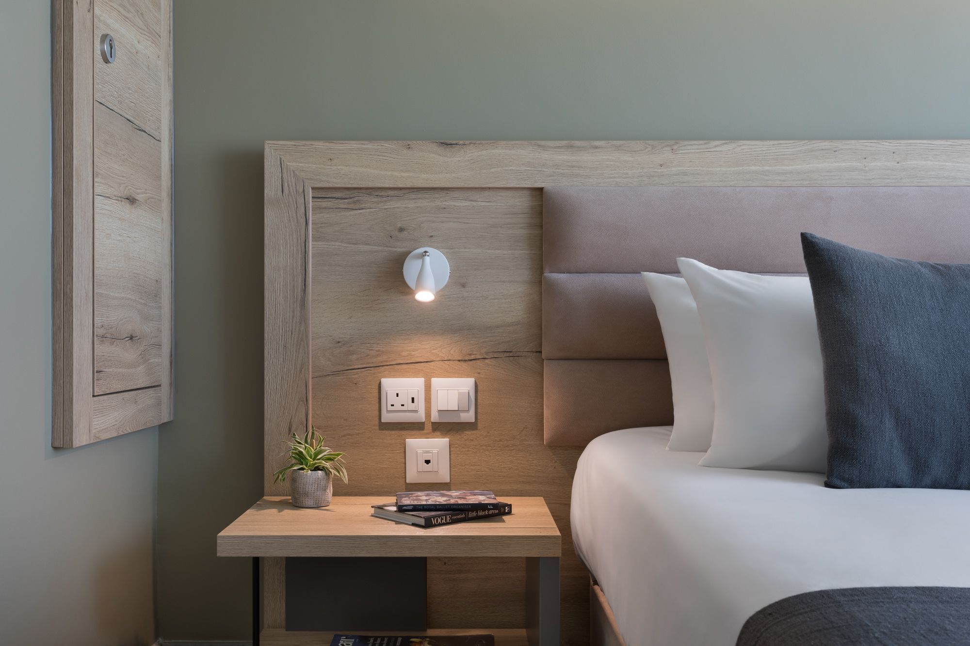 A neatly arranged hotel room with a wooden headboard, a bedside table with a plant and books, and a bed with pillows.