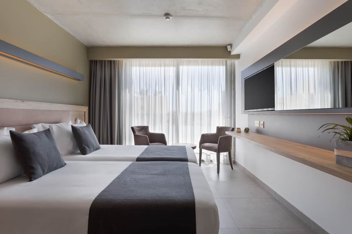A modern hotel room with two beds, a TV, a desk, and a large window with curtains.
