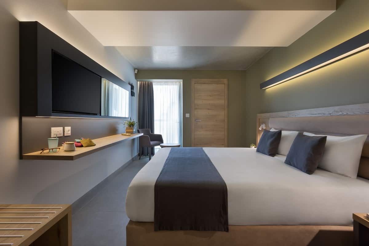 A modern hotel room with a large bed, a flat-screen TV mounted on the wall, a bedside table with a lamp, and a window with curtains.