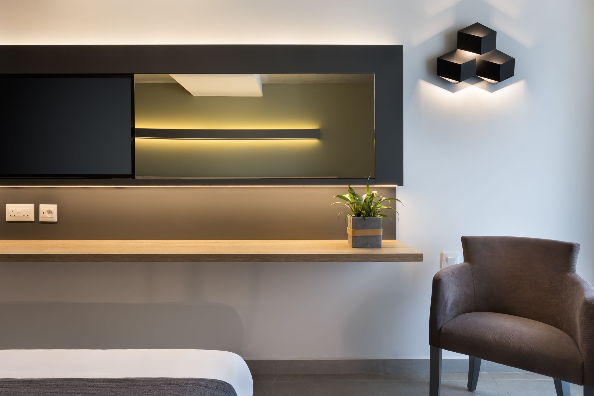 A modern hotel room with a large flat-screen TV mounted on the wall, a wooden shelf with a plant, and a brown armchair.