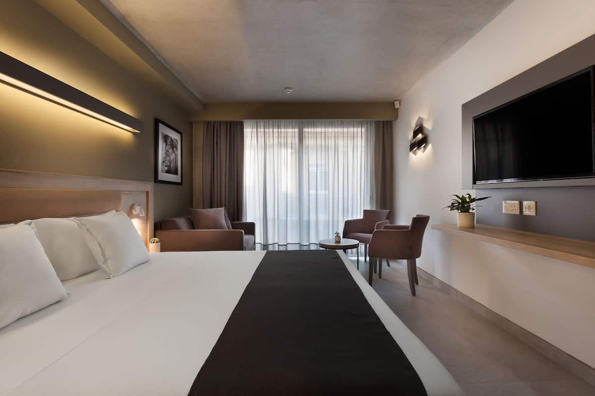 A hotel room with a large bed, a TV mounted on the wall, a desk with a plant, and a view of the city through the window.