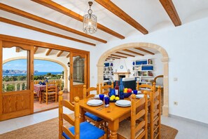 Un coin repas avec une table en bois dressée pour quatre personnes, une vue sur la mer par une porte ouverte et une bibliothèque en arrière-plan.