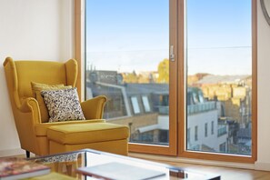 A yellow armchair with a patterned pillow next to a large window with a city view.