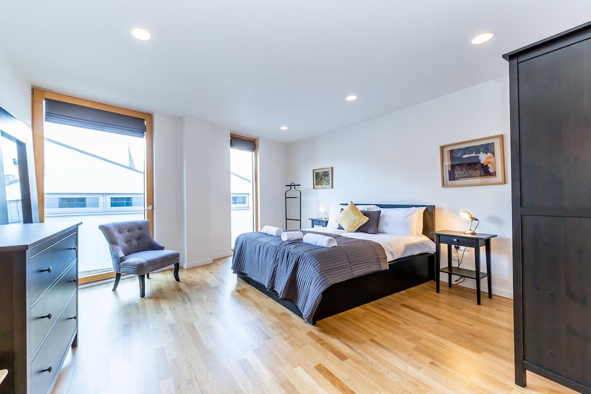 Een moderne slaapkamer met een bed, nachtkastje, stoel, dressoir en een groot raam.