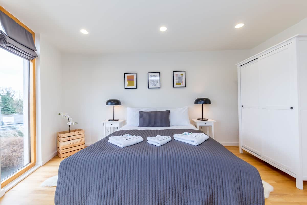 Een moderne slaapkamer met een groot bed, twee nachtkastjes met lampen, een garderobe en een raam met gordijnen.