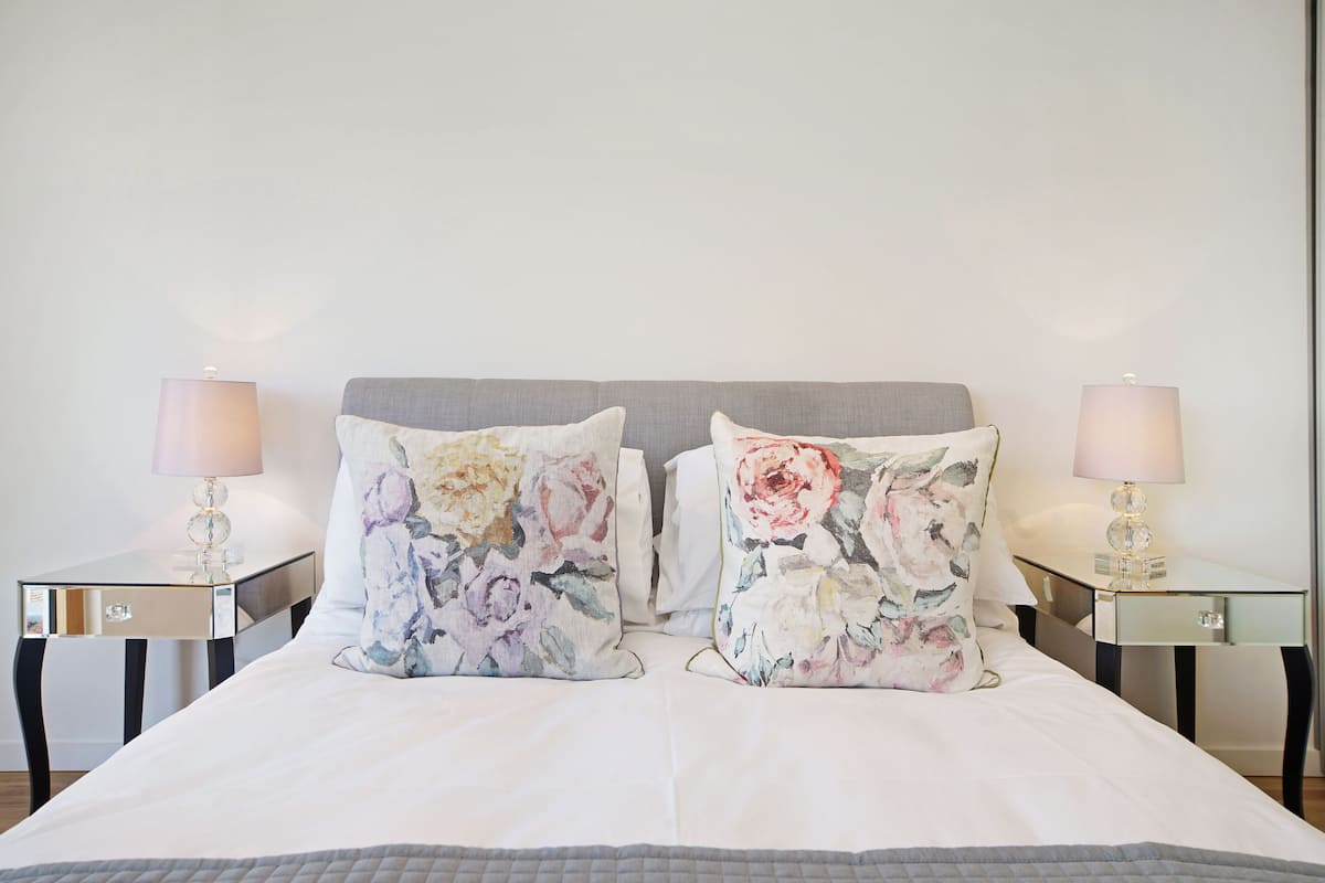 Een net opgemaakt bed met twee bloemenkussens, een grijs hoofdbord en twee nachtkastjes met lampen.