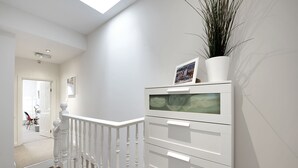 A white staircase with a railing, a white wall with a picture frame, and a plant on top of a white cabinet. - Finding My Palace in Tooting (London)