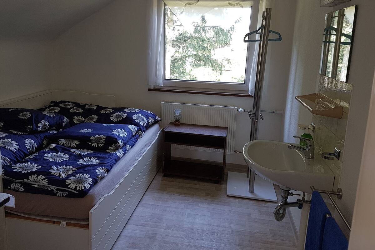 Ein Schlafzimmer mit Bett, Fenster, Heizkörper, Nachttisch und einem Badezimmer mit Waschbecken und Spiegel.
