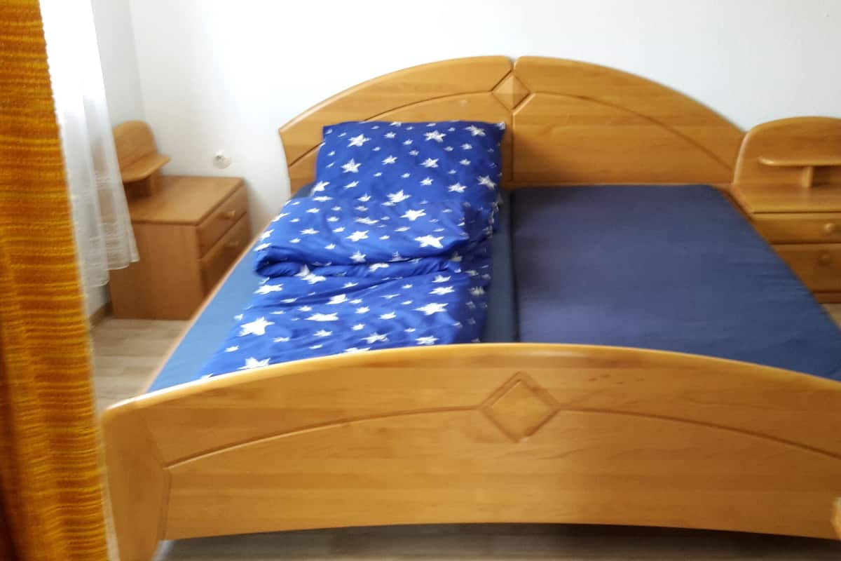 Ein Einzelbett mit Holz-Kopfstück, einer blauen Matratze mit weißen Sternen und einem passenden Kissen. Am Bett steht ein Nachttisch mit einer Lampe.
