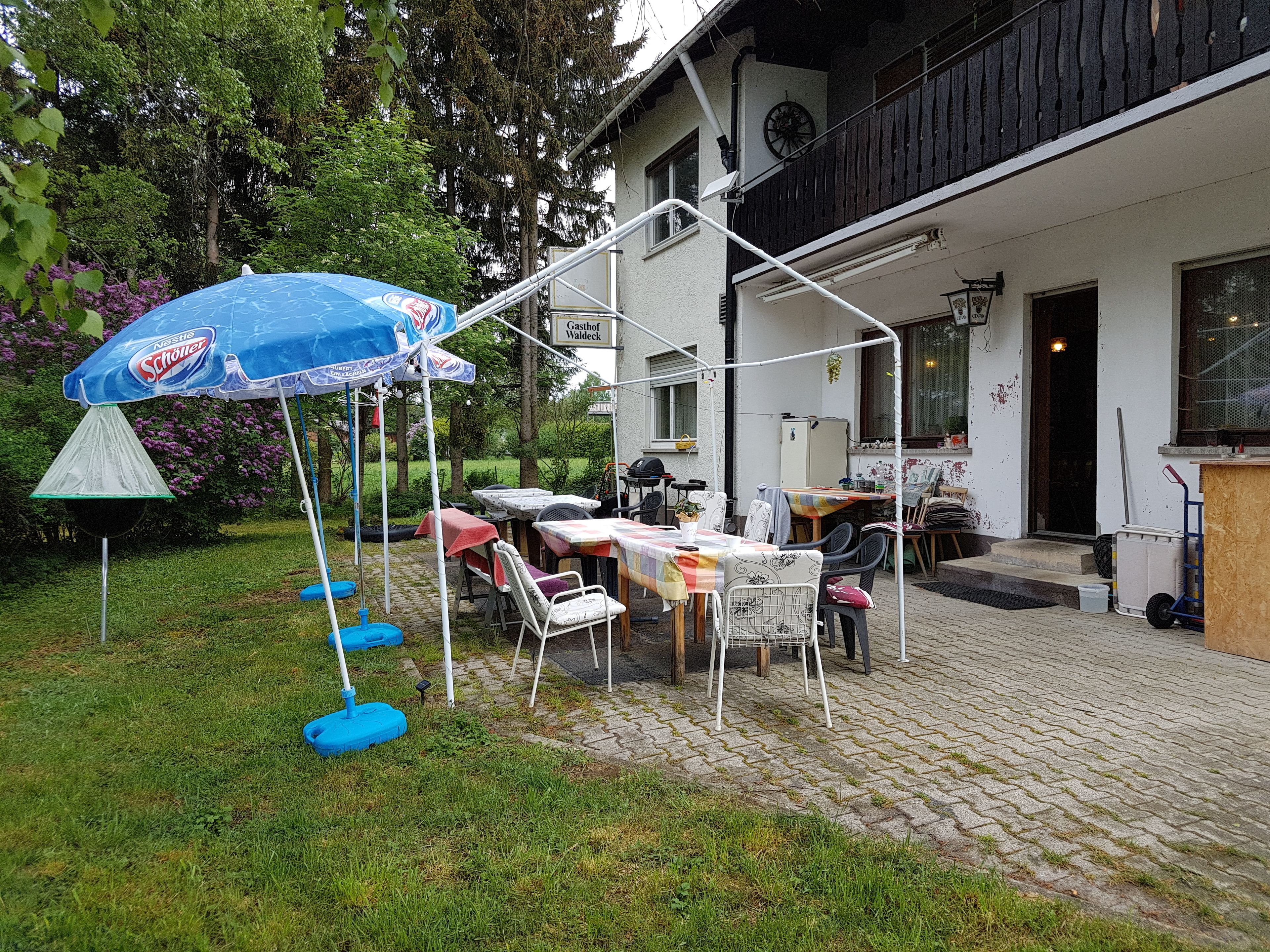 An outdoor patio area with a table and chairs, a blue umbrella, and a garden setting.